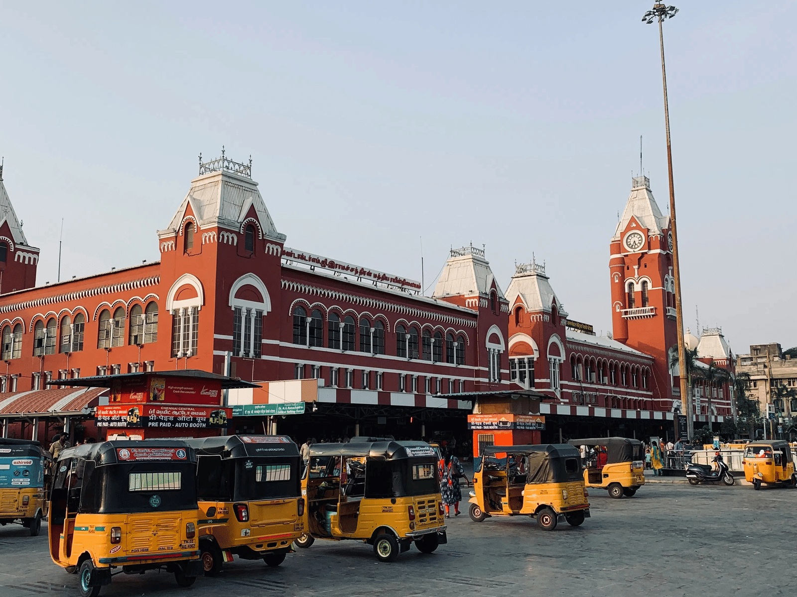 The iconic red Gothic-style facade of Chennai Central Railway Station, featuring its grand clock tower and several yellow auto-rickshaws parked in the foreground.