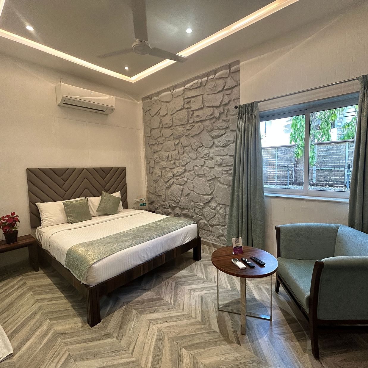 A bedroom featuring a plush king size bed, elegant stone-textured walls, and a cosy seating area, illuminated by natural light streaming through large windows – Maxxvalue Klub Klass Villa, Lonavala.