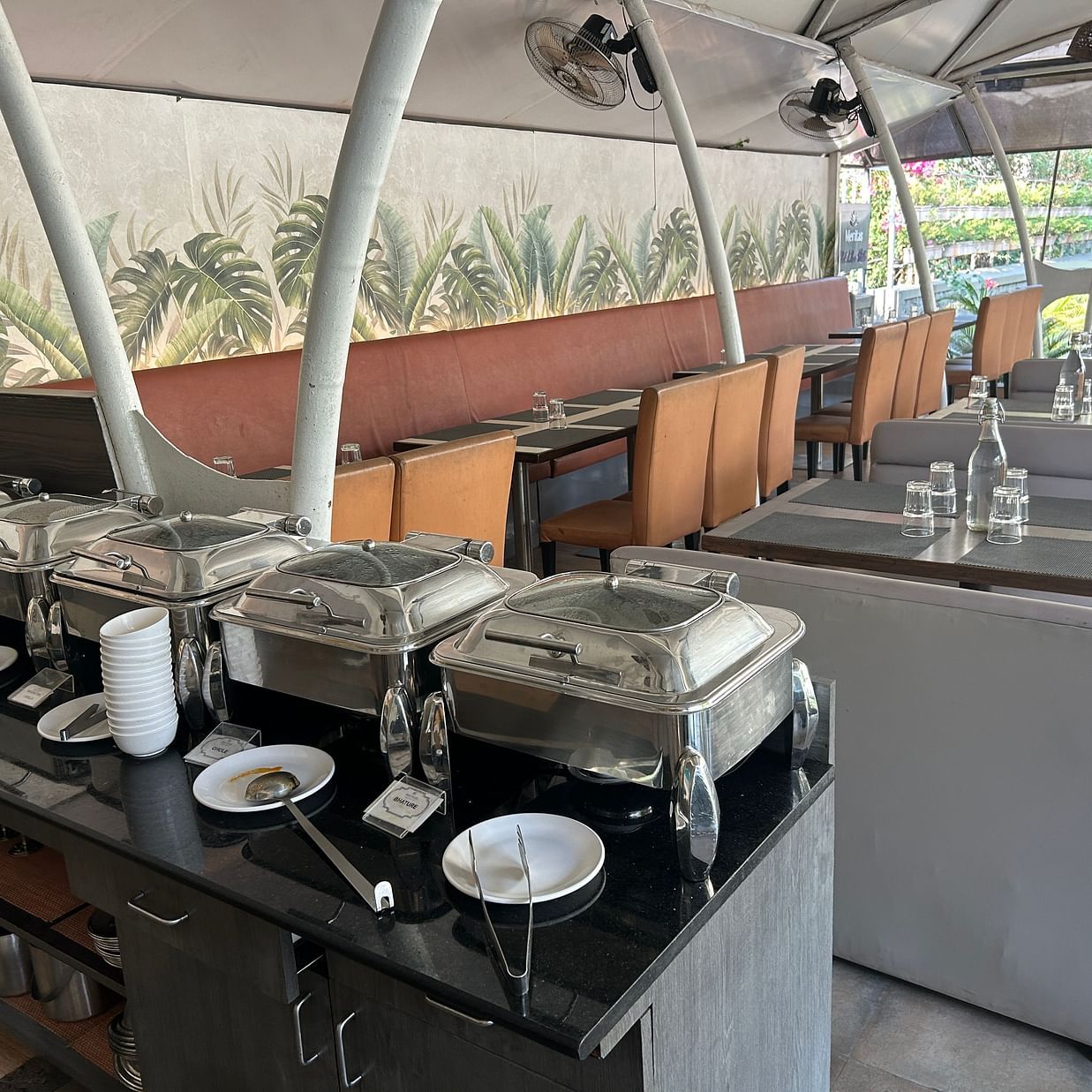 A well-arranged buffet dining setup with stainless steel chafing dishes, plates, and cutlery, set in a semi-outdoor setting– Maxxvalue Klub Klass Villa, Lonavala.