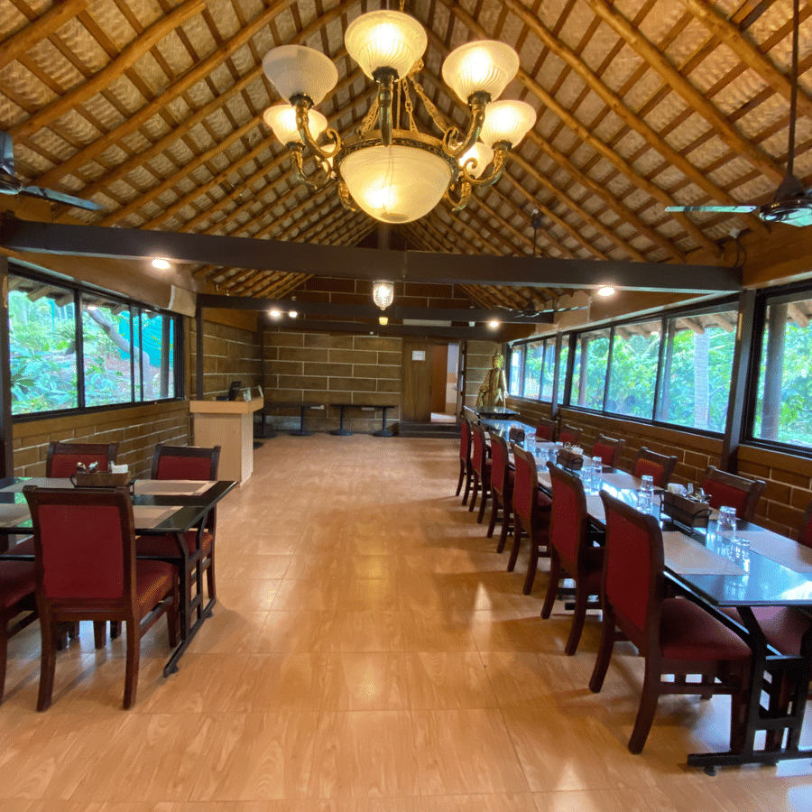Formal dining setup at Ibex River Resort, Pollachi showcasing wooden tables and chairs, elegant chandeliers, and large windows providing natural light and scenic forested views for guests.