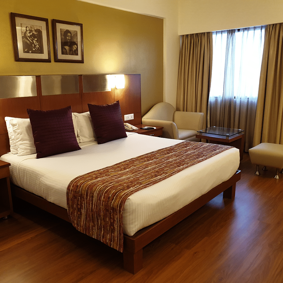 An image of a Deluxe Room with double size bed, bedside table and lam on either side and a huge window covered with curtains - The Suryaa New Delhi