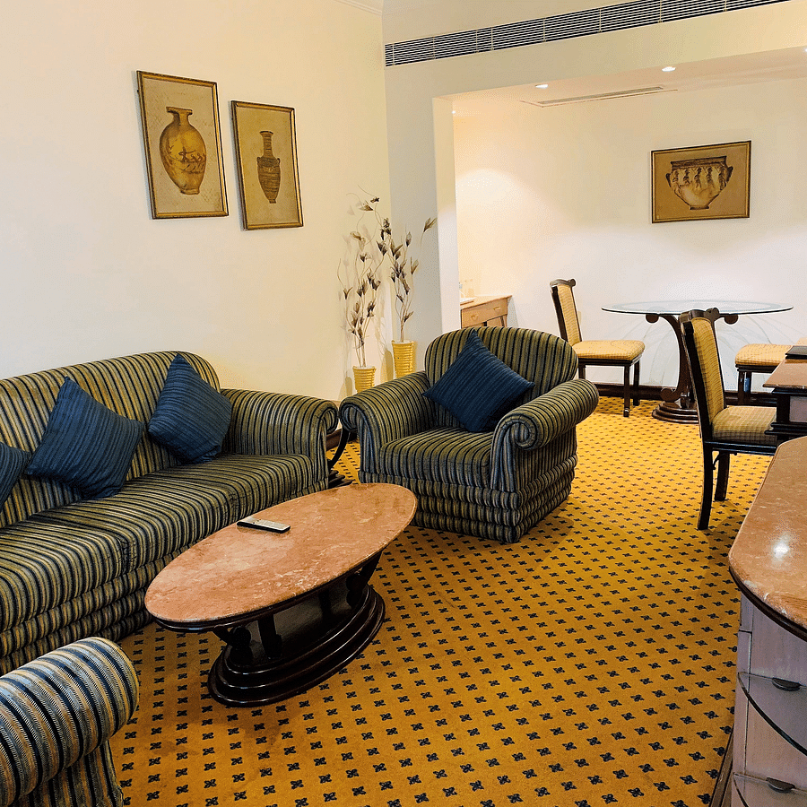 An image of a seating area with sofa sets, centre table, flat TV screen, and a cabinet in a Deluxe Suite - The Suryaa New Delhi