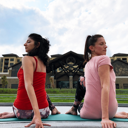 2 women seated back-to-back on a yoga mat outdoors, performing a twisting pose with the facade of YO1 Longevity and Health Resorts in the background.