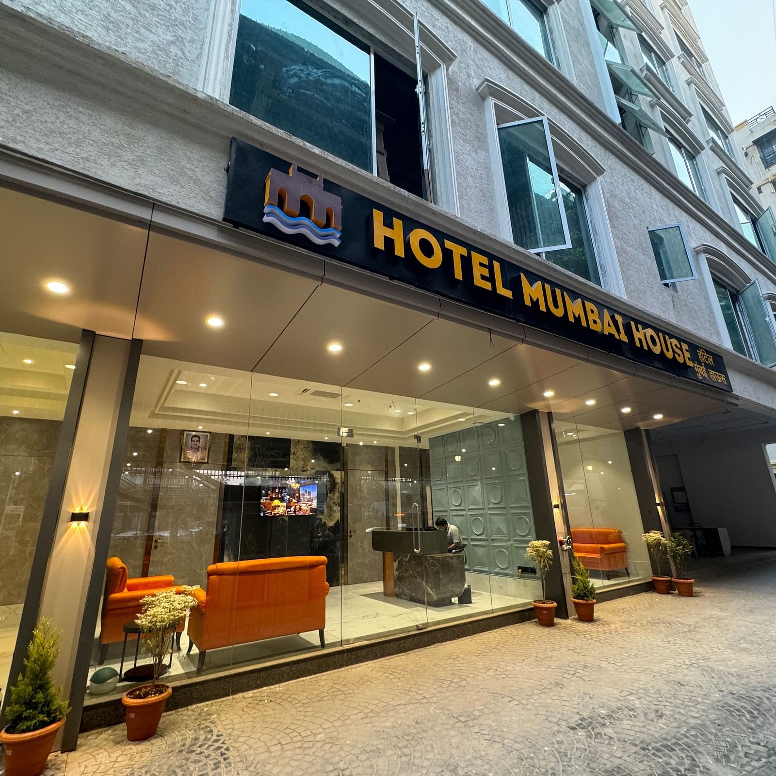 The entrance of Hotel Mumbai House with a modern design, showcasing a sign above the door, glass walls, a sofa set in the foyer, and potted plants on the sidewalk.