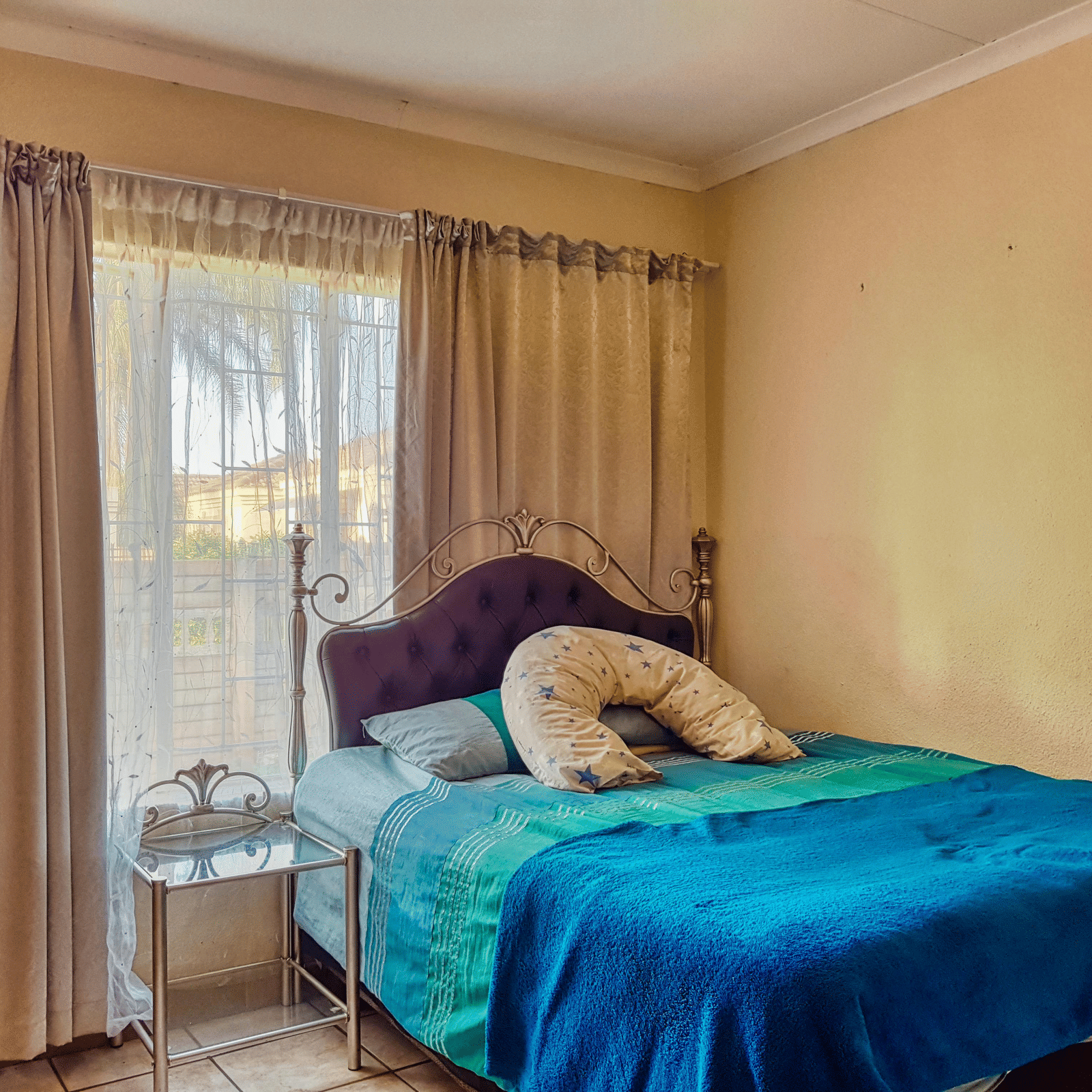A bedroom featuring a double bed with sheets spread over it with beige curtains covering the window behind it.