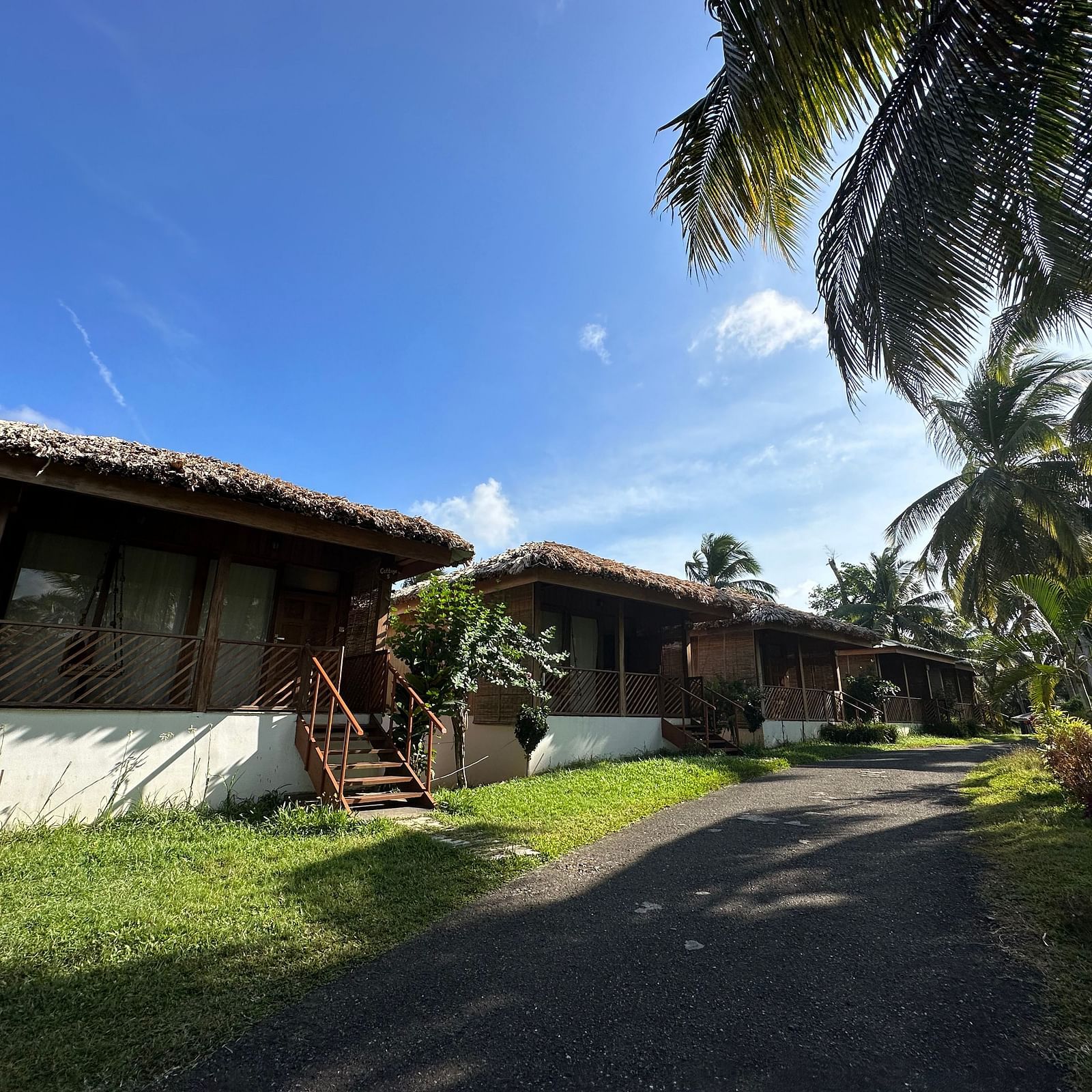 A charming resort building with a thatched roof, surrounded by lush green grass and palm trees under a bright sky.