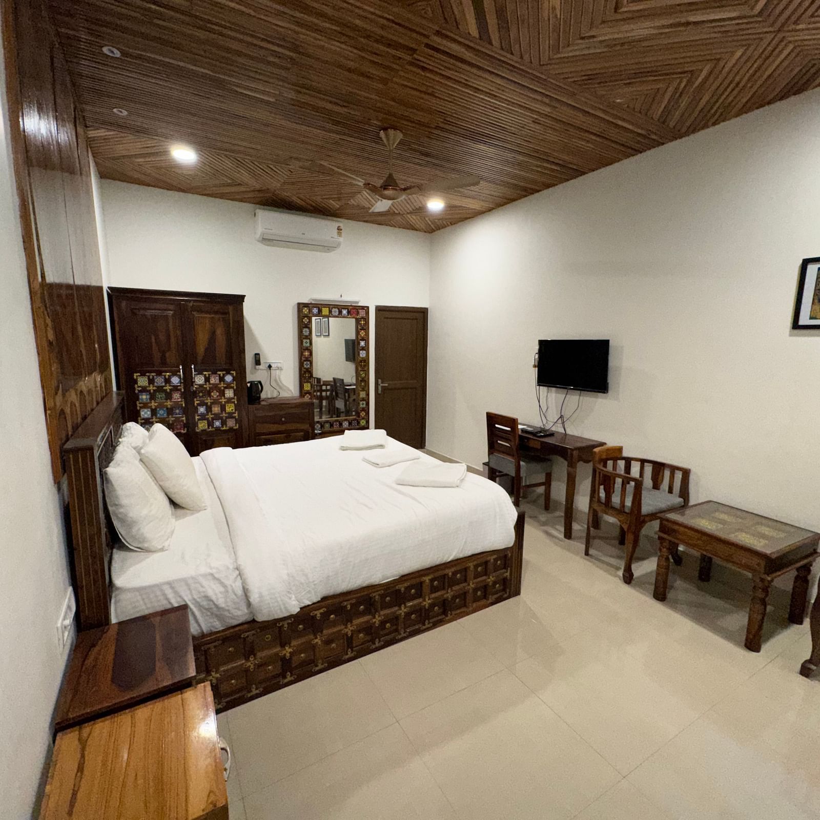 A hotel bedroom featuring a large white bed, dark wooden furniture including a TV cabinet, and a textured wooden ceiling.