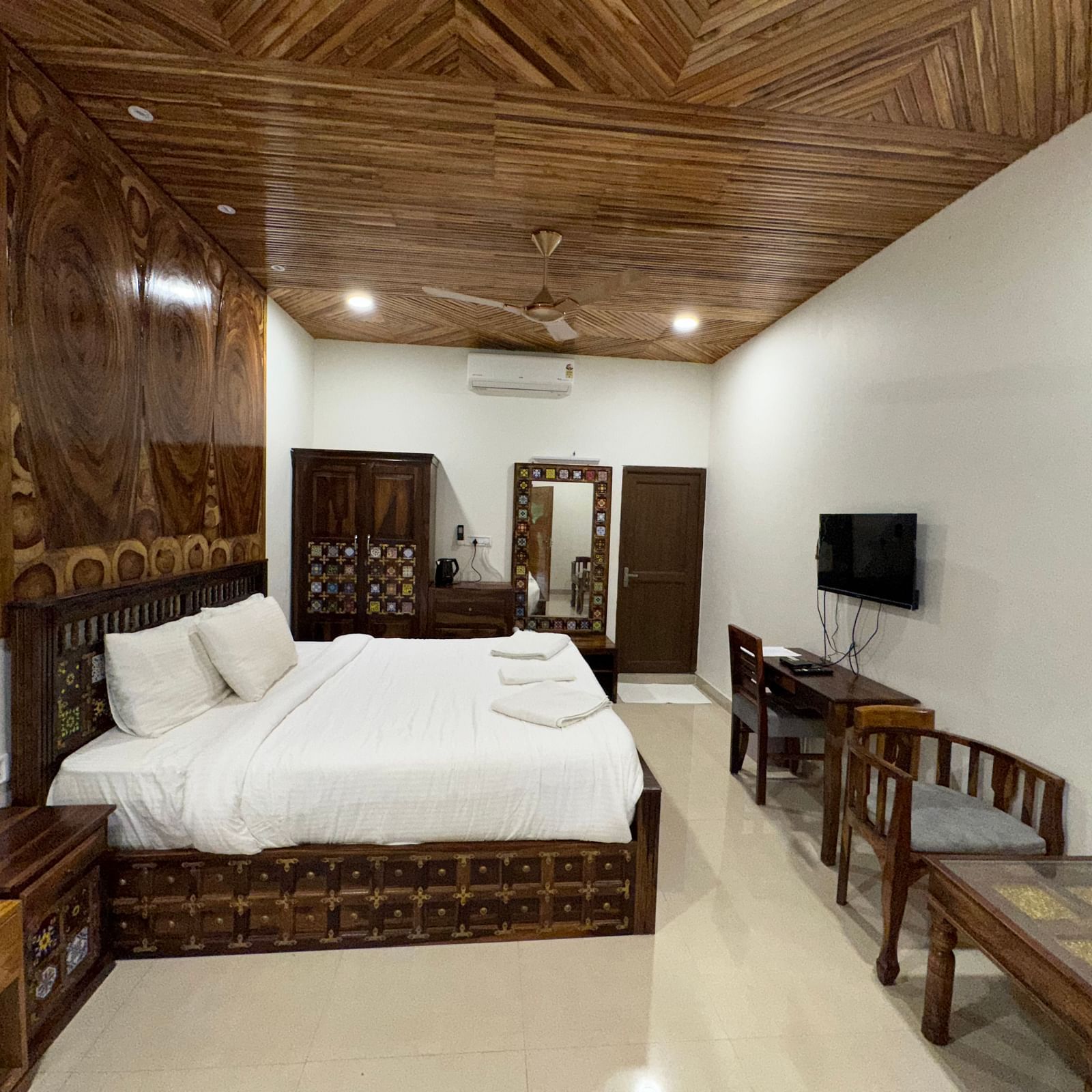 A hotel bedroom with a large bed, wooden furniture, a TV, and a unique patterned wooden ceiling.
