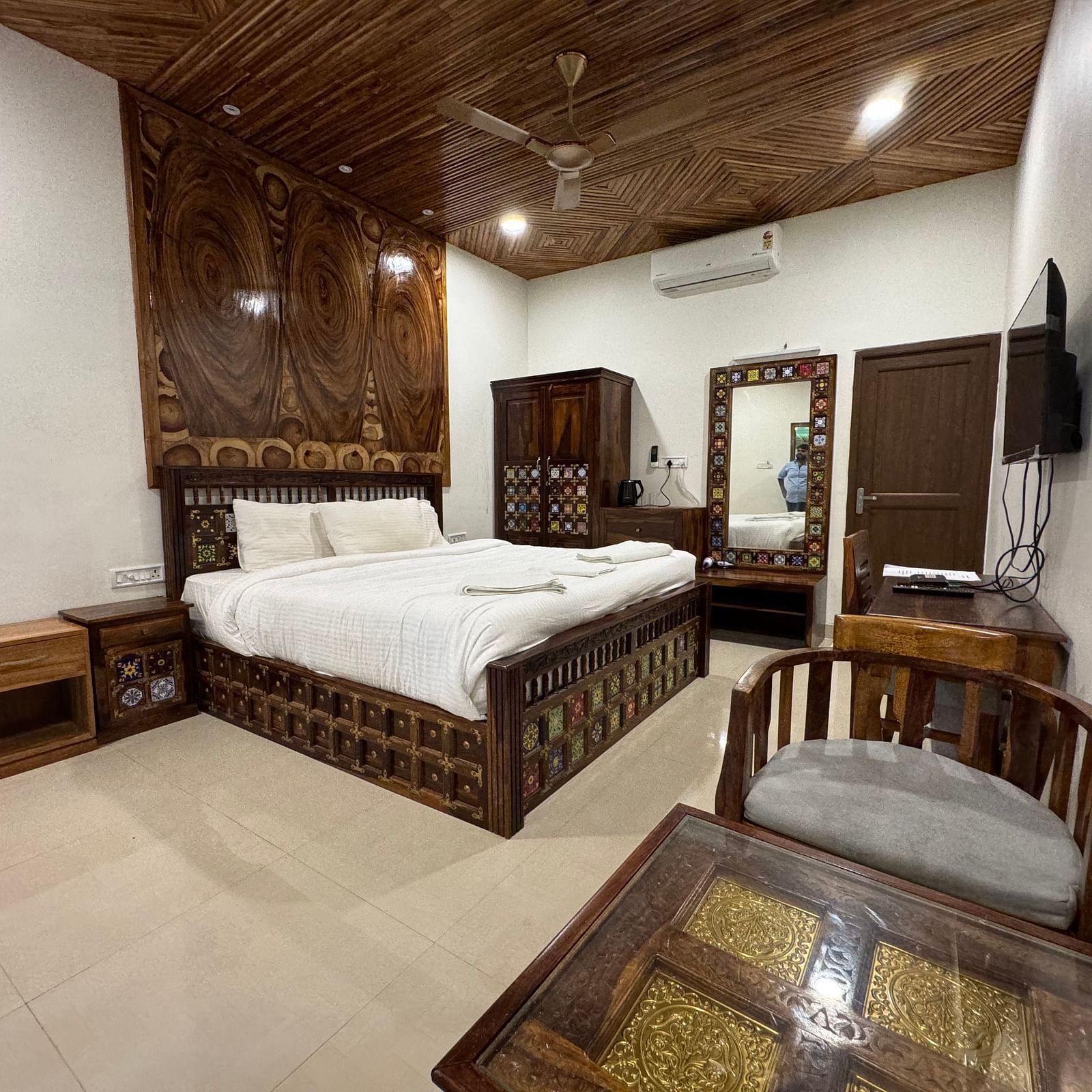 A hotel bedroom with a prominent bed, matching wooden nightstands, a dressing table, and a decorative wooden ceiling.