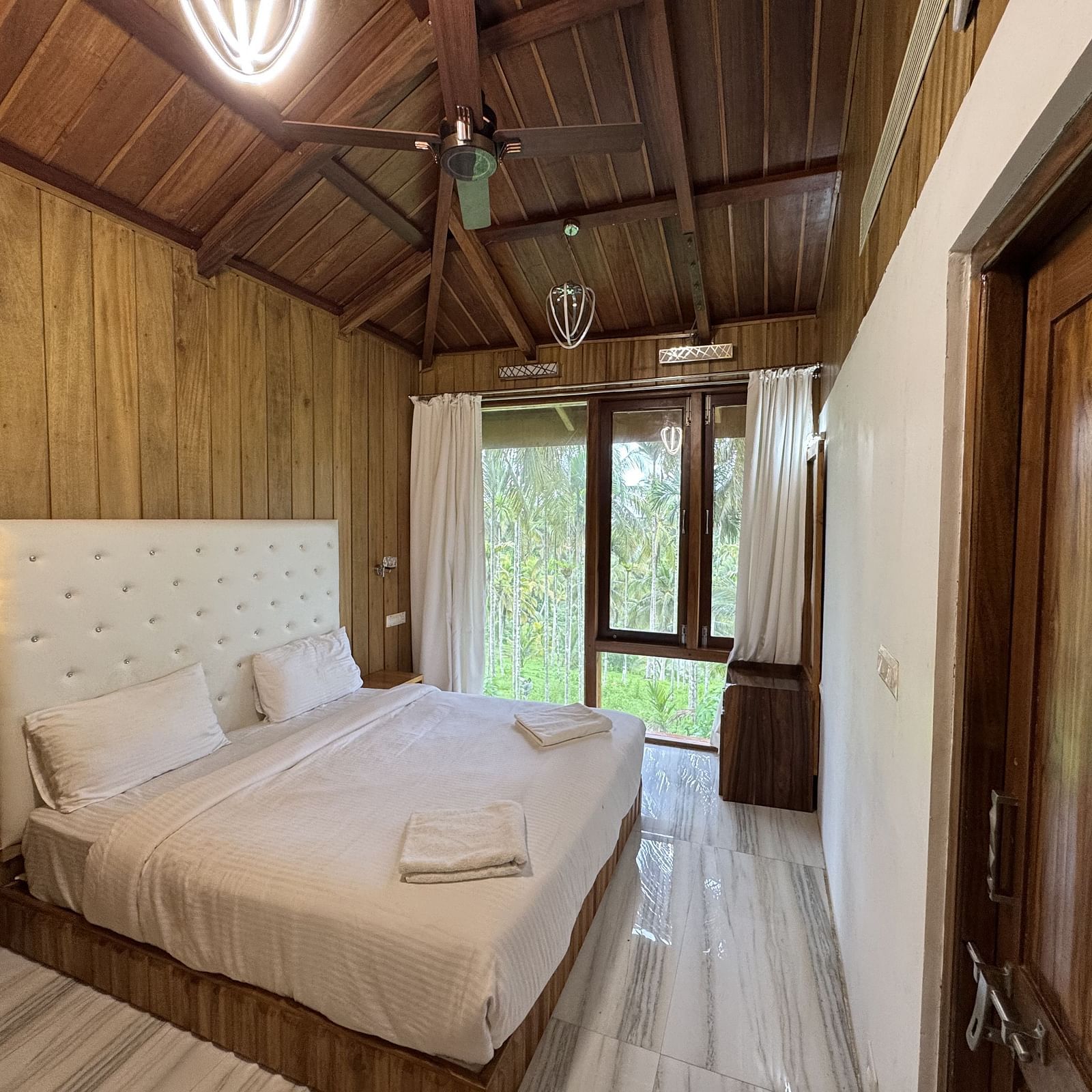A resort bedroom with two beds, wooden walls and ceiling, a ceiling fan, and large windows with curtains.