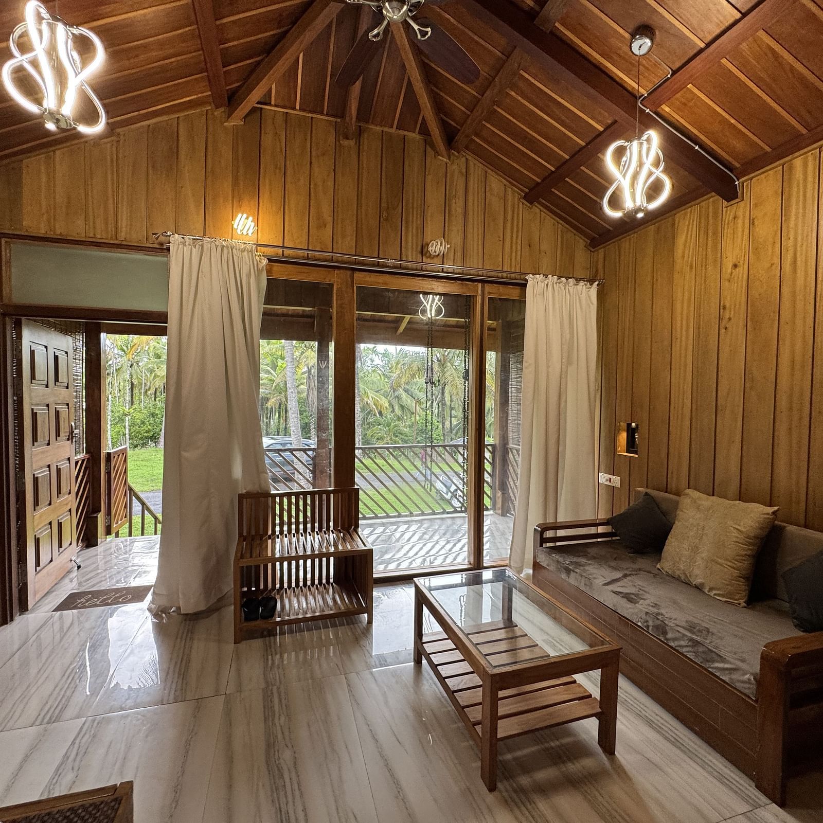 A rustic living room with wooden walls and ceiling, a sofa, a coffee table, and open doors leading to an outdoor view.