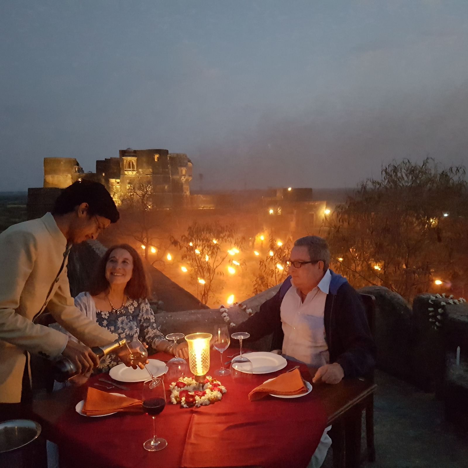 A waiter serves a meal to two people at a candlelit table on a rooftop at dusk, with lights visible in the distance- Shahpura Bagh.