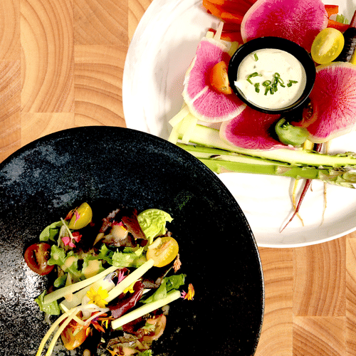 2 artfully plated gourmet salads with fresh vegetables and dipping sauce, served on black and white plates over a wooden table surface at YO1