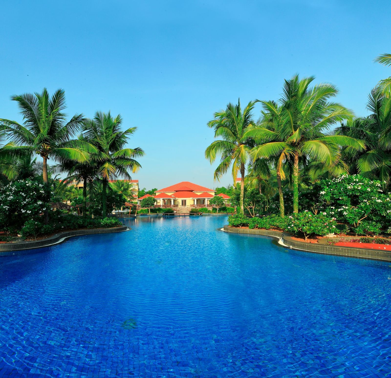 A beautiful swimming pool at a tropical resort, surrounded by palm trees and lush greenery and a building with a red roof in the distance - Ocean Spray, Pondicherry