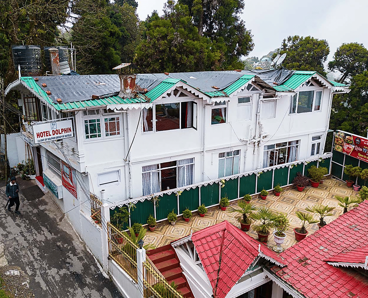 An aerial view of a multi-level white hotel building with green and red roofs nestled among trees on a hillside at Hotel Dolphin Darjeeling.