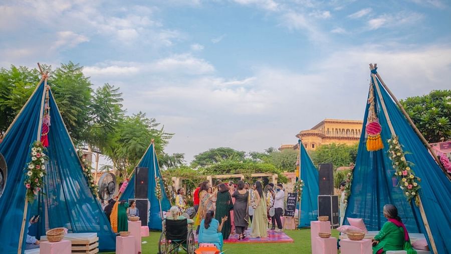 Blue tents and seating are arranged on a green lawn for an event at Tijara Fort-Palace - 19th Century, Alwar under a partly cloudy sky.