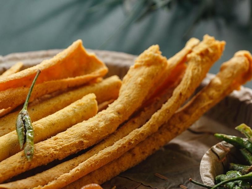 Golden, crispy fried sticks arranged on a plate with fried green chili garnishes. Background is softly blurred, highlighting the crunchy texture of the snacks.