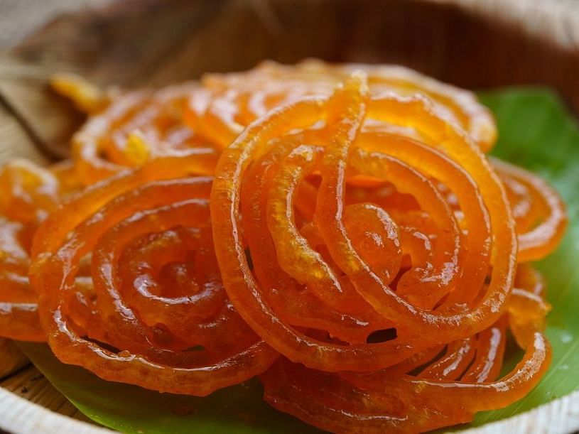 Close-up of  jalebi, a spiral-shaped Indian dessert, resting on a leaf on a wooden plate. The jalebi is glossy and appears syrup-coated, showcasing its intricate loops .
