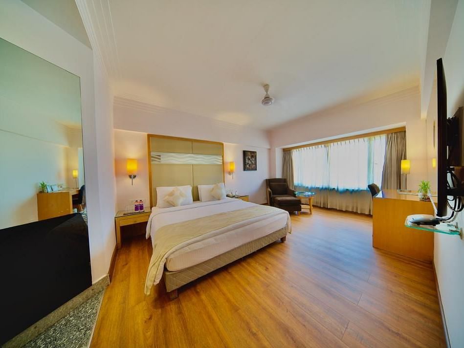 An elegant bedroom in Lords Plaza Ankleshwar with a double bed, wooden panelled floor, large mirror, TV, dim light lamps, and ample natural light from a large window.