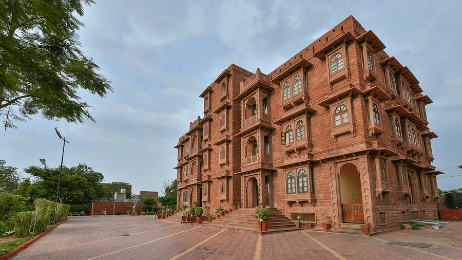 A multi-storey stone building at Garh Govind standing next to a paved courtyard under a cloudy sky with trees to the left.