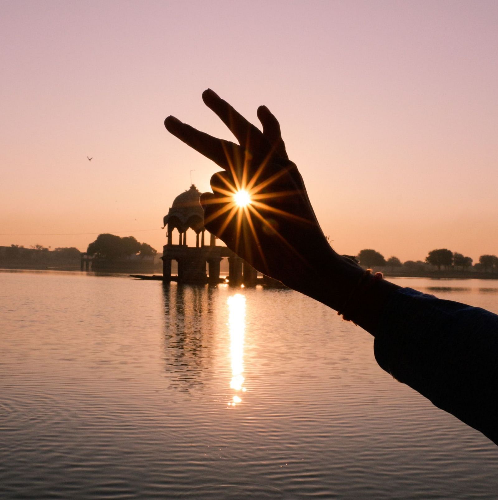 Silhouette of a hand framing the sunrise over a lake with a pavilion in the background.
