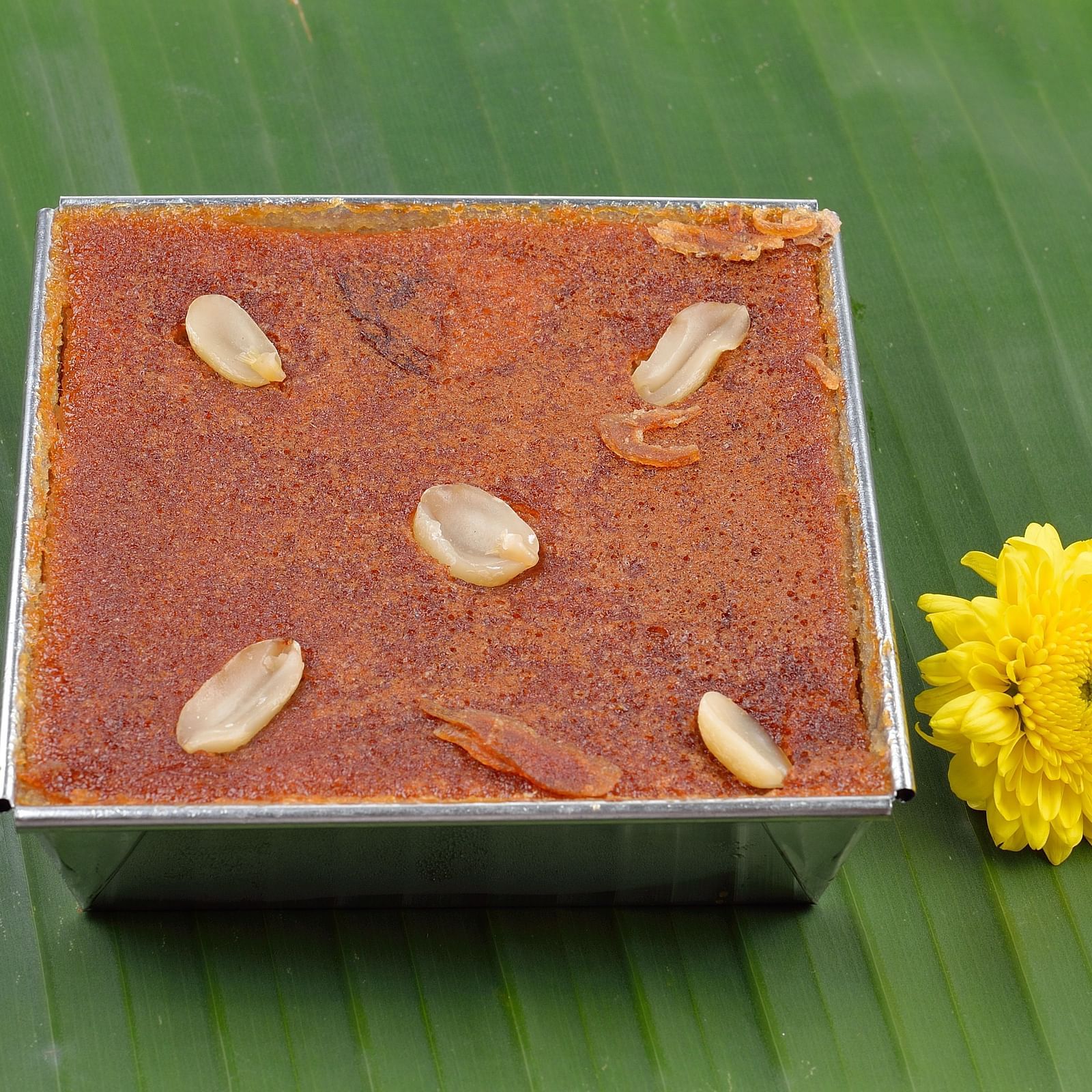 A square tin of a sweet, rich Indian dessert, garnished with nuts and a yellow flower.