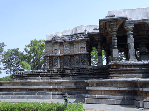 A side view of the Hoysaleswara Temple, Halebidu, with intricate carvings on the walls and pillars, and trees in the background.