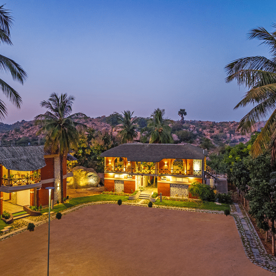 The resort buildings illuminated at dusk, showcasing the red-brick architecture and sandy courtyard - Nature Trails Ashoka Resort Hampi