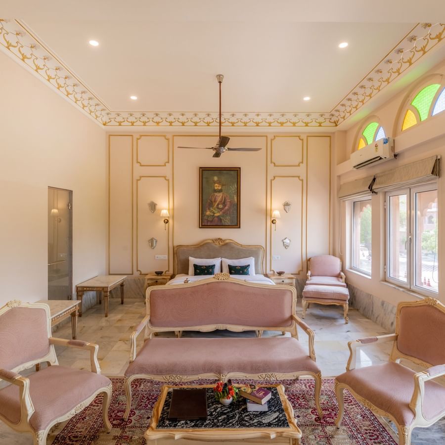 Front view of Maharaja Suite at Umaid Palace featuring a comfortable sitting area with sofa set and centre table. There is a bed behind the sofa with lamps on wither side, and two windows.
