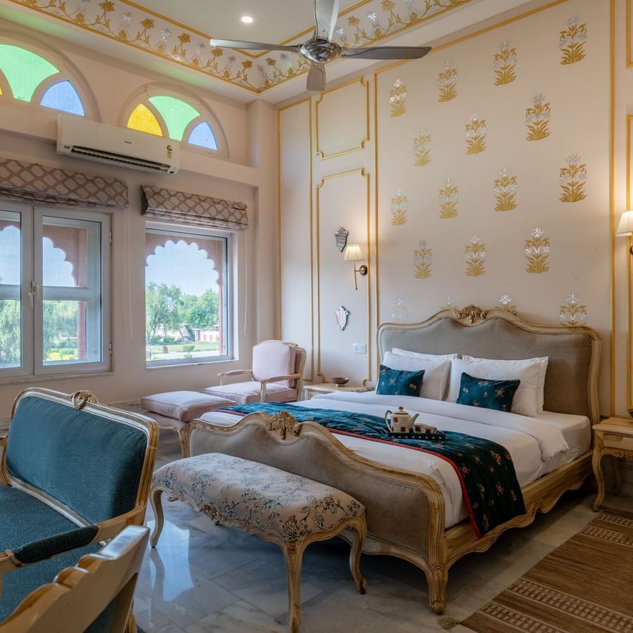 Maharaja Suite at Umaid Palace featuring a lavish bedroom with king size bedside table and lamps on either side. The room also have few soft sofa chairs an, a table, and a window near AC.