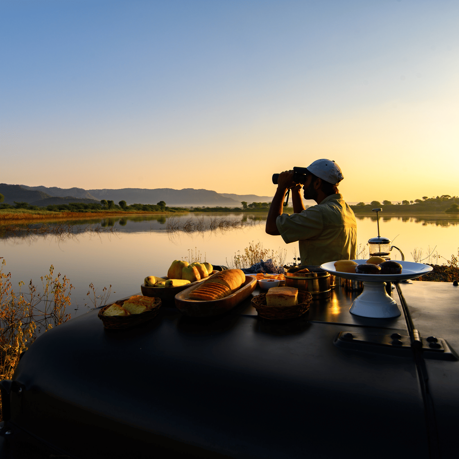 An image of a man standing near a lake with his jeep parked over looking the mountain - An image of a jeep parked amidst the rocky mountains - Utsav Camp Sariska