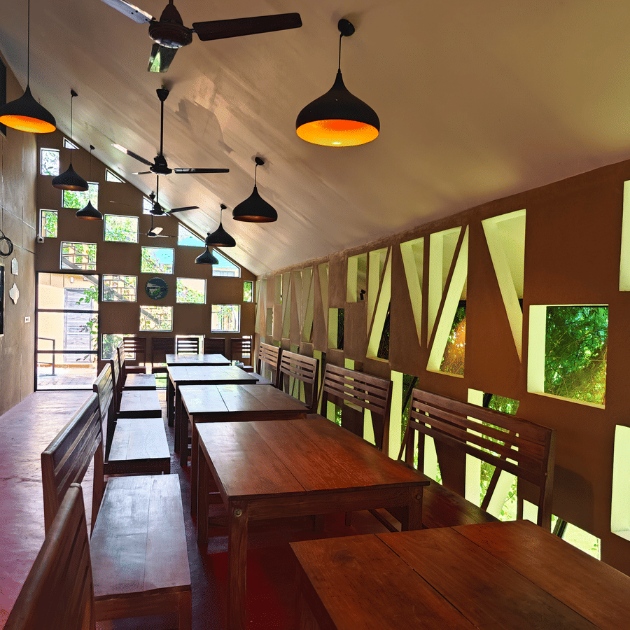 Interior of a rustic dining hall with long wooden tables, warm pendant lighting, and a unique geometric window wall - Ibex Resorts, Malampuzha (Kava Eco Camp and Caravan Park)