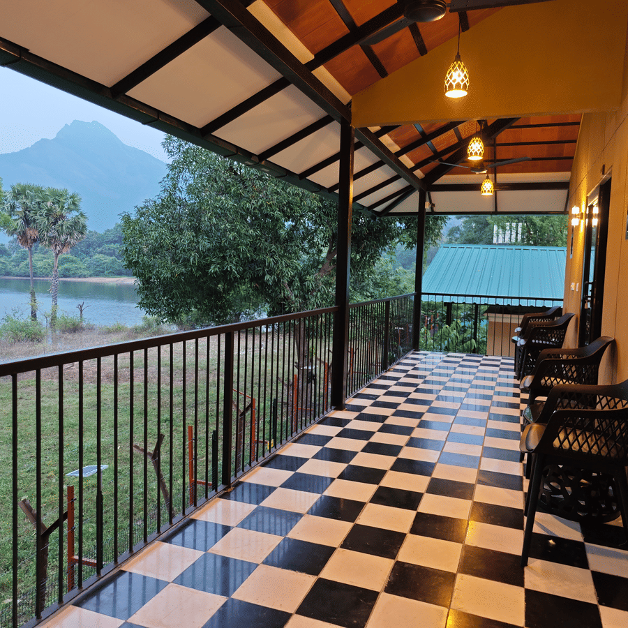 A long veranda with black and white checker flooring and seating, offering a view of mountains and a lake - Ibex Resorts, Malampuzha (Kava Eco Camp and Caravan Park)