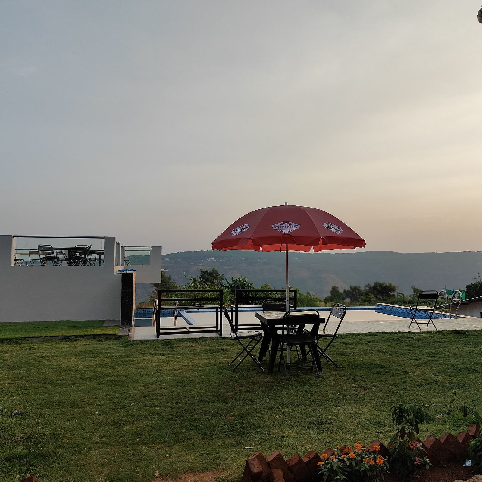 Outdoor area featuring a red umbrella and seating area on a green lawn and a swimming pool, with hills visible in the background.