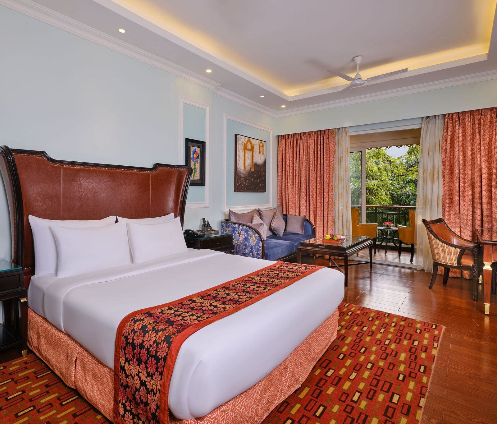 elegant Executive Room with a large bed, rug, tables, chairs, paintings on walls and large window with curtains overlooking greenery