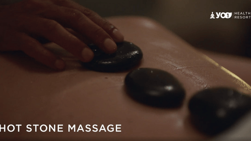 A person's back during a hot stone massage, with several dark, smooth stones placed along the spine and a hand adjusting one at YO1 Longevity & Health Resorts