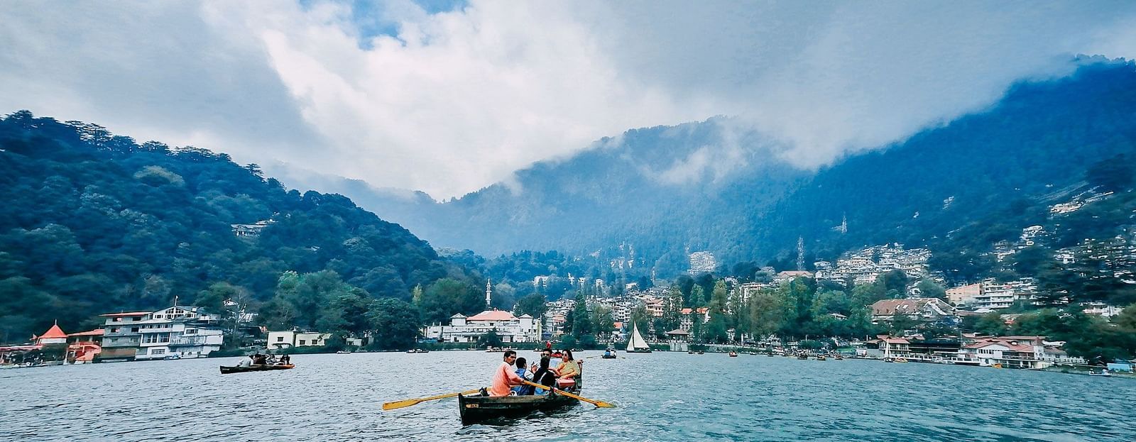 Tourists boating on the tranquil blue waters of Nainital Lake, with forested hills and scattered buildings in the background under a misty sky.
