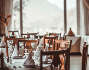 A restaurant dining area with wooden chairs, white tablecloths, folded napkins, and large windows.