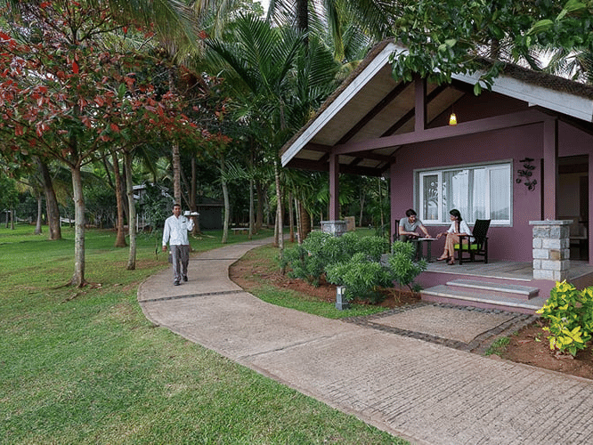 Waterfront Villa at The Serai Kabini featuring a staff member walking on the pathway surrounded by verdant manicured lawn and a couple of guests on the patio