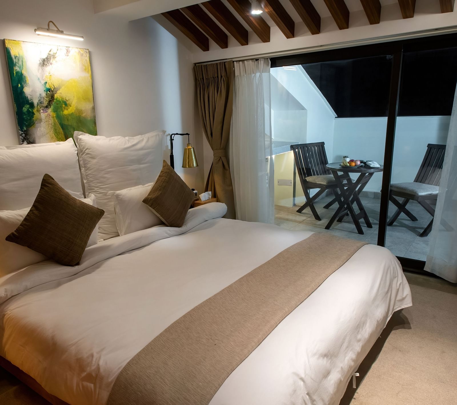 Loft suite featuring a bed with pillows and cushions with a painting on the wall and an expansive glass door leading to the balcony where chairs and table are placed - The Nanee 