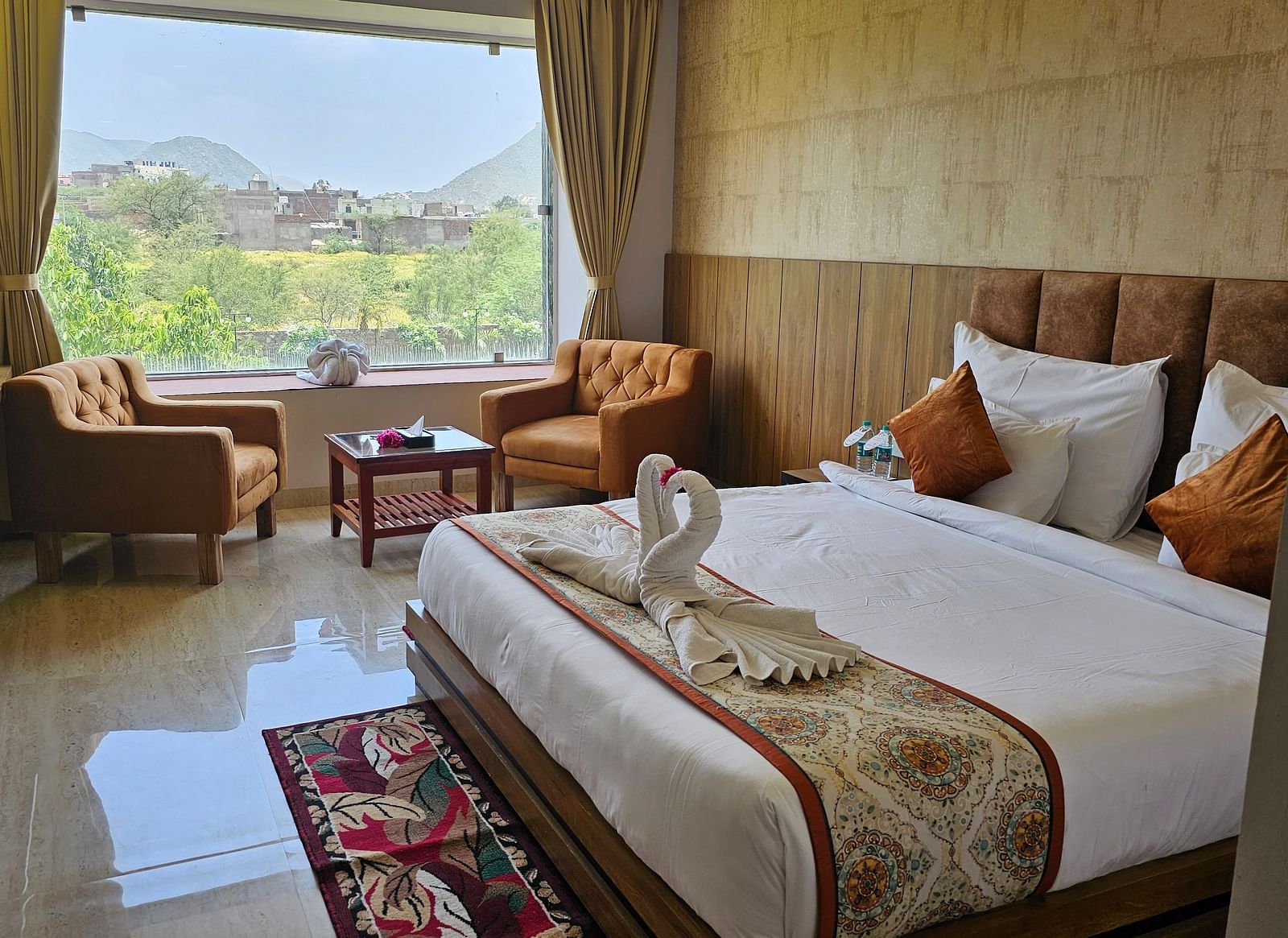 A bright and spacious resort room at Bamboo Saa Mulberry Resort, Pushkar, featuring a large window with a view of the outdoors including a made bed with a patterned runner, two armchairs, and a small table.