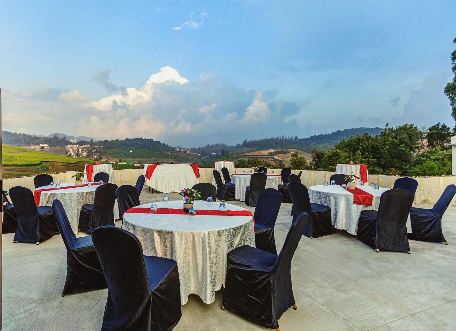 Rooftop event setup at Mango Hill Shola, Ooty, featuring round tables covered in white linens with red and black accents, surrounded by black banquet chairs, overlooking lush green hills and a dramatic cloudy sky.