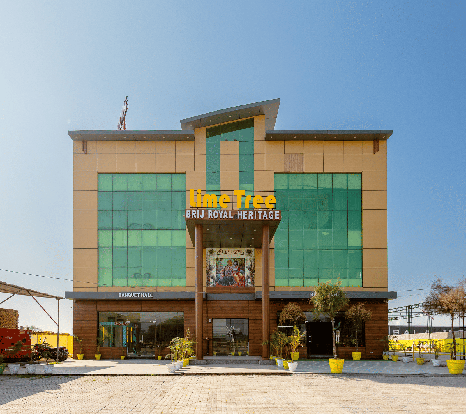 A daytime view of Lime Tree Hotels & Banquet, Vrindavan building with glass panels, yellow accents, and an open parking area in front.