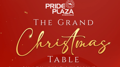 A banner of The Grand Christmas Table dinner buffet and music event at Pride Plaza Ahmedabad featuring a curtain backdrop, gold script lettering, 2 large christmas trees, a central plate of plated food, and small desserts and cocktails in the foreground.