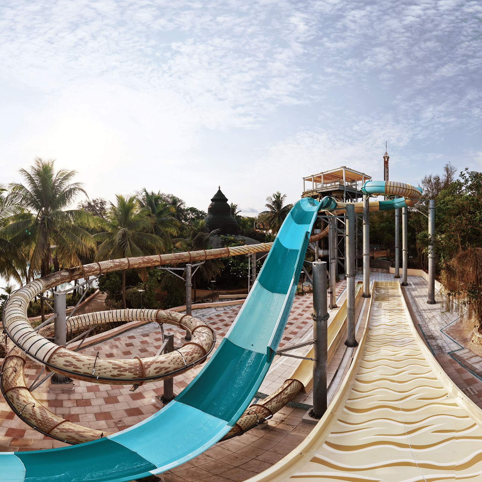 The blue and white open-air water slide of the Abyss Plunge ride, twisting downward alongside a beige exit chute, surrounded by palm trees and lush greenery at Water Kingdom.