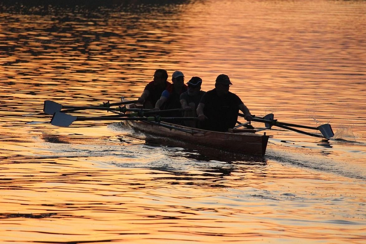 People rowing a boat with oars over a lake reflecting a golden sunset.