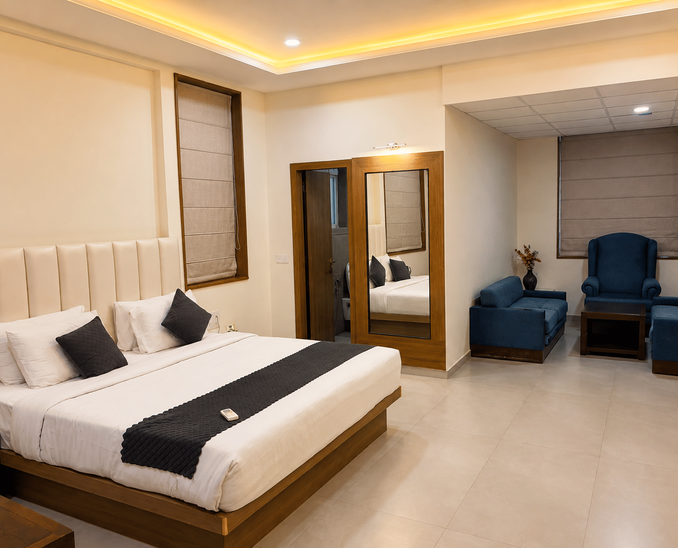 Suite at TGI Apple Inn, Ajmer Road, Jaipur featuring a bed with pillows, a seating area with armchairs and a table, blinds on the windows, and a doorway.
