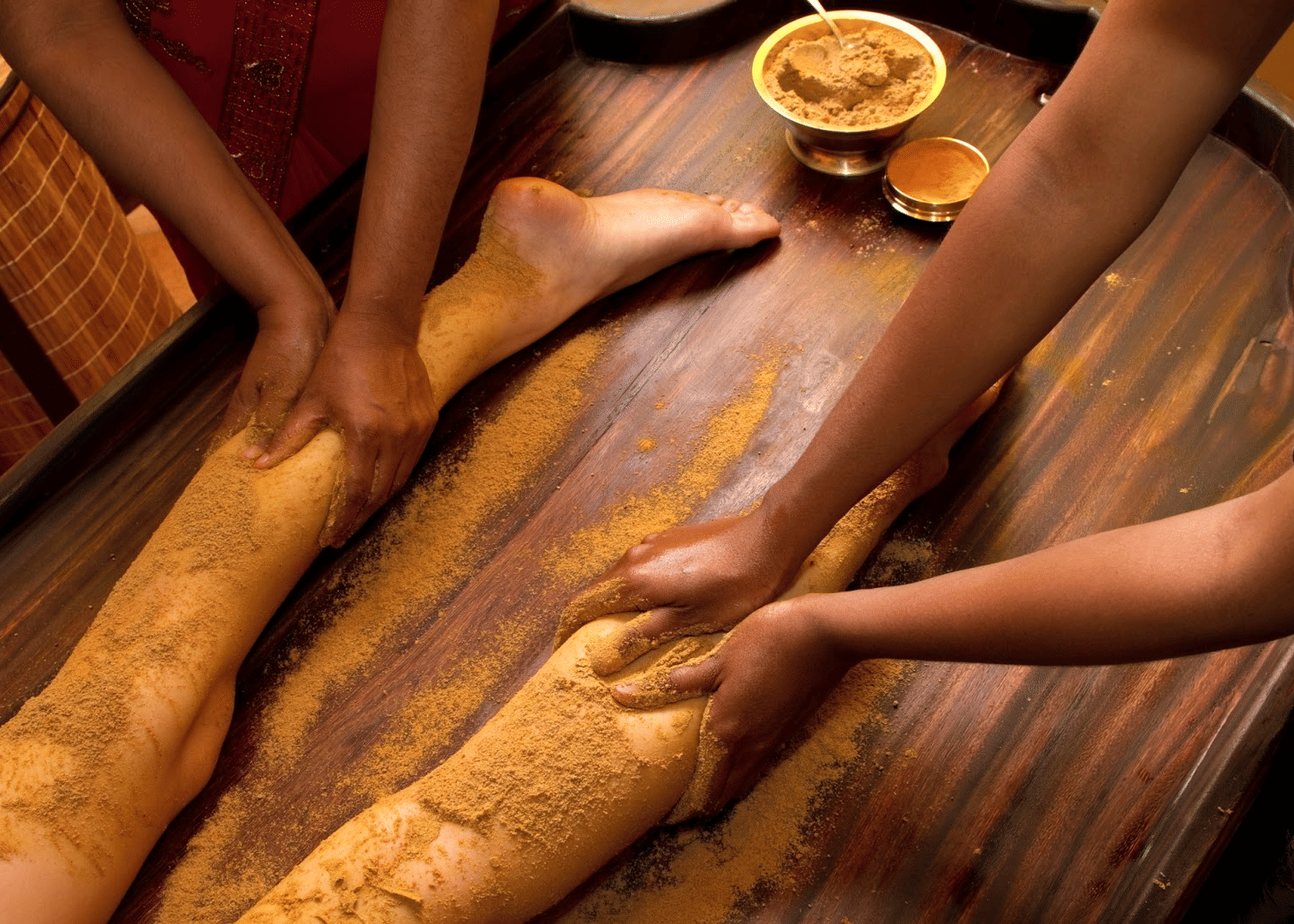 Close-up of hands applying herbal paste on a person's legs during a massage, with a wooden surface and bowl of paste visible at Paradise Lagoon Resort, Udupi.