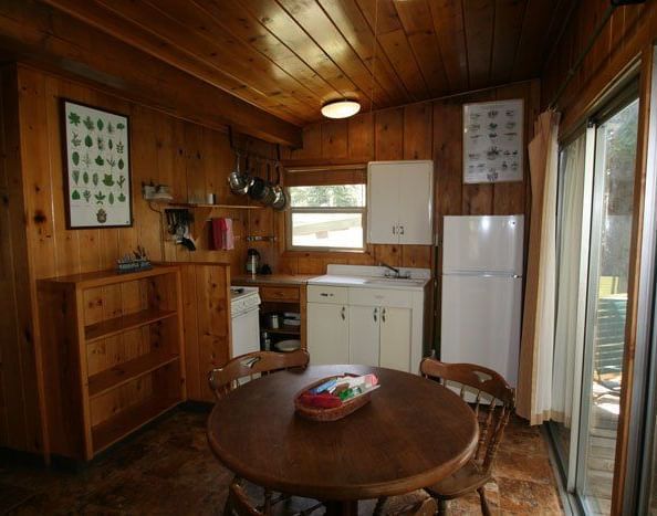 24The interior of a cabin showing a dining area with a round wooden table and chairs, adjacent to a kitchen area