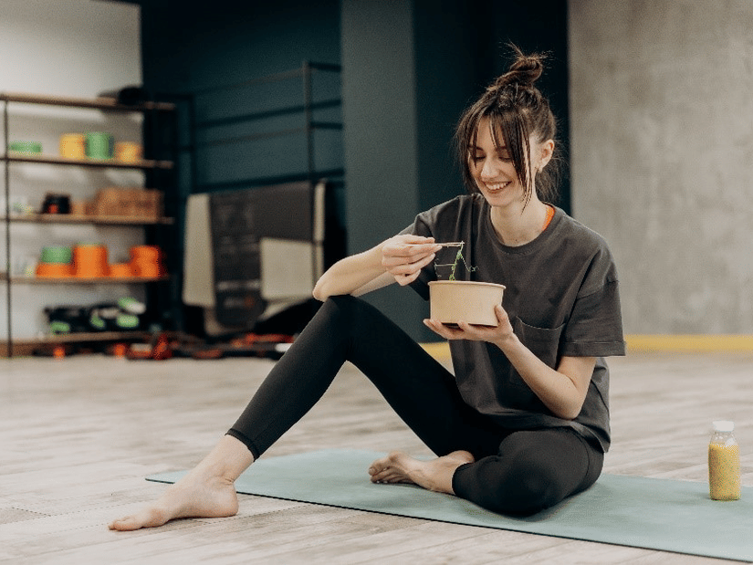A woman sits barefoot on a yoga mat in a fitness studio, smiling while enjoying a healthy meal with a juice bottle beside her.